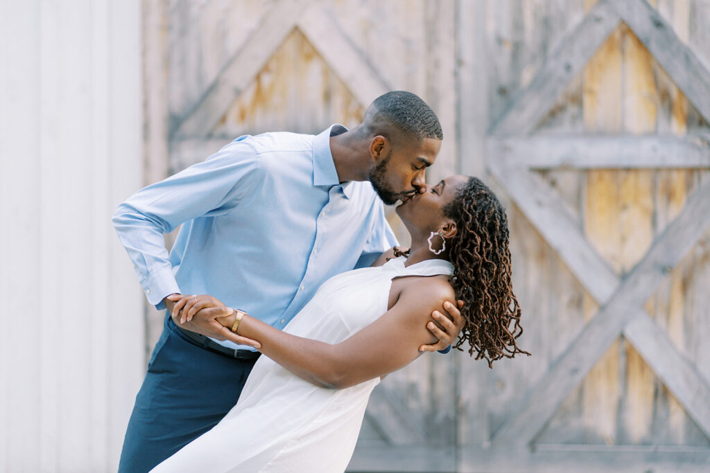 A close-up of Agnes and Uzo in front of the rustic barn doors at Union Grove Barn in Chapel Hill, North Carolina. Agnes wears a chic white jumpsuit while Uzo pairs a blue button-down with khakis, adding stylish touches to their engagement session, captured by North Carolina wedding photographer Jessica Leigh Mason.
