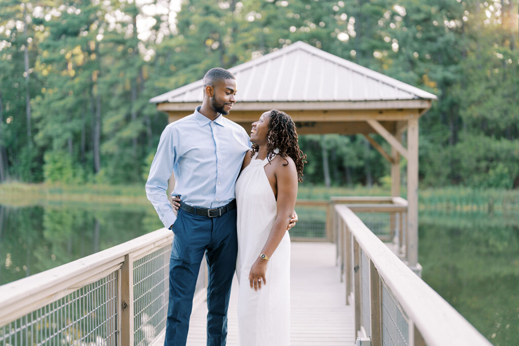 A wide view of Agnes and Uzo standing on the walkway by the lake at Union Grove Barn in Chapel Hill, North Carolina, sharing a sweet moment looking at each other during their engagement session, captured by North Carolina wedding photographer Jessica Leigh Mason.