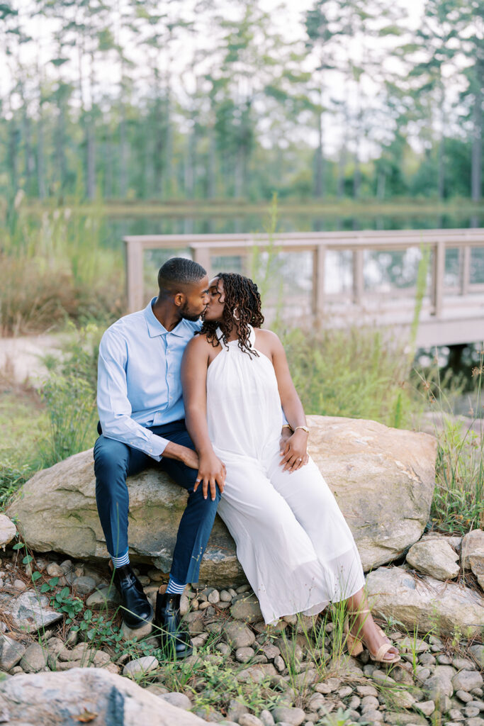 Agnes and Uzo sit together on a boulder by the water’s edge at Union Grove Barn in Chapel Hill, North Carolina, sharing a peaceful moment during their engagement session, captured by Durham wedding photographer  Jessica Leigh Mason.