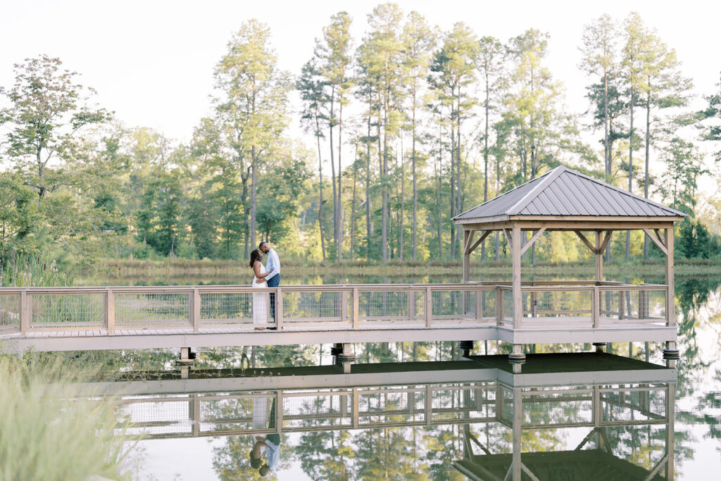 Agnes and Uzo stand on the walkway leading to the gazebo beside a mirror-like lake at Union Grove Barn in Chapel Hill, North Carolina, capturing a romantic summer engagement with true NC countryside charm, documented by North Carolina wedding photographer Jessica Leigh Mason.