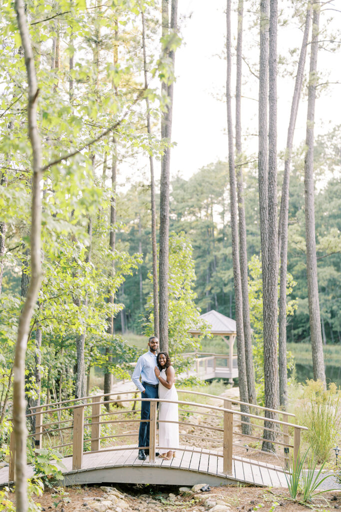 Agnes and Uzo stand close together on the bridge above a gentle creek, surrounded by towering pines at Union Grove Barn in Chapel Hill, North Carolina, captured by North Carolina wedding photographer Jessica Leigh Mason.