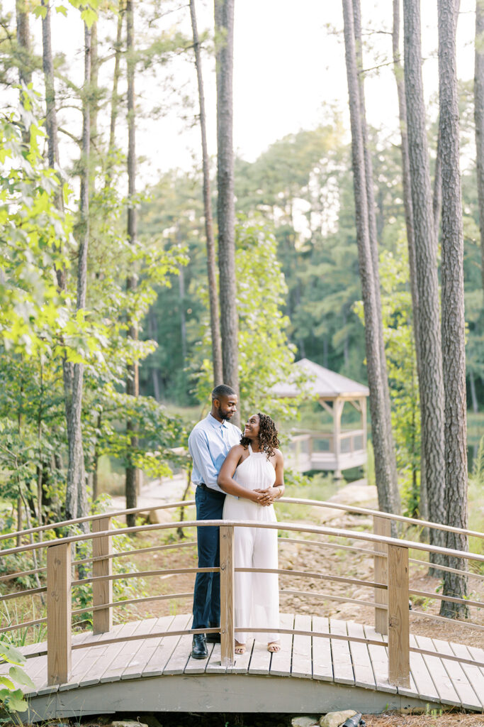Agnes and Uzo stand together on the bridge over a babbling creek, surrounded by beautiful tall pines at Union Grove Barn in Chapel Hill, North Carolina, captured by North Carolina wedding photographer Jessica Leigh Mason.