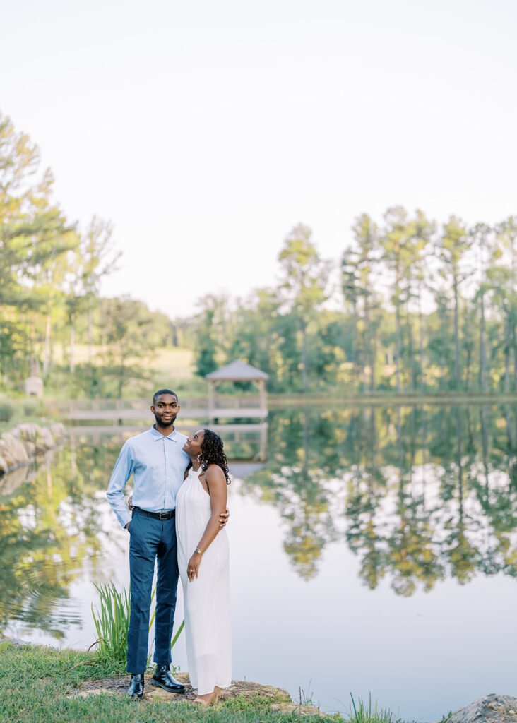 Agnes and Uzo stand near the still, mirror-like lake reflecting the trees and sky at Union Grove Barn in Chapel Hill, North Carolina, captured by North Carolina wedding photographer Jessica Leigh Mason.