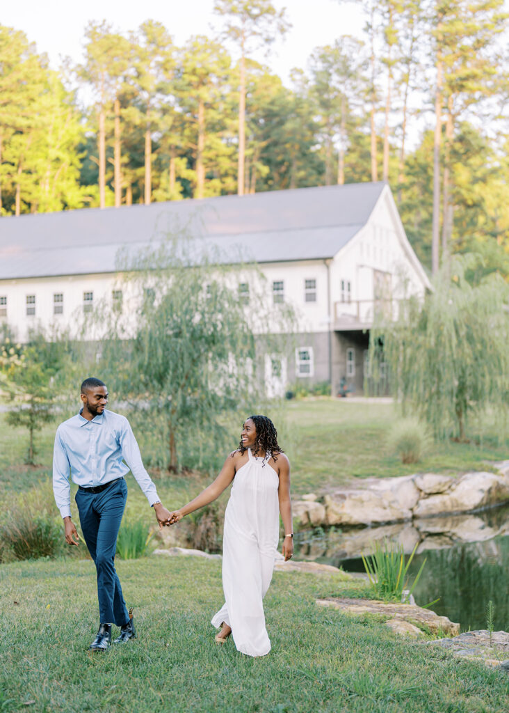 Agnes and Uzo walk hand in hand along the lake with the beautiful wedding barn in the background at Union Grove Barn in Chapel Hill, North Carolina, captured by North Carolina wedding photographer Jessica Leigh Mason.