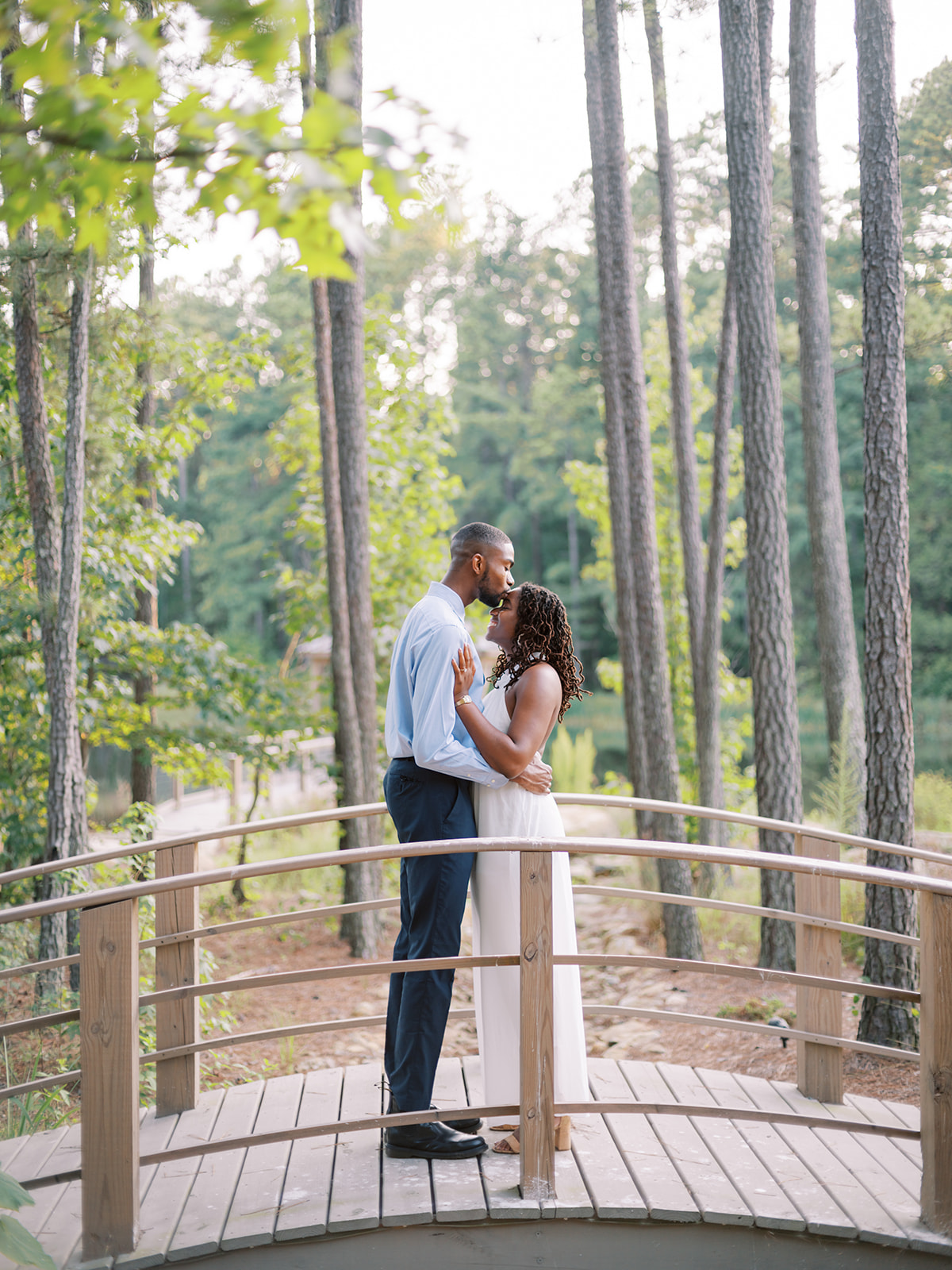 Agnes and Uzo stand close together on the bridge above a gentle creek, surrounded by towering pines at Union Grove Barn in Chapel Hill, North Carolina, captured by North Carolina wedding photographer Jessica Leigh Mason.