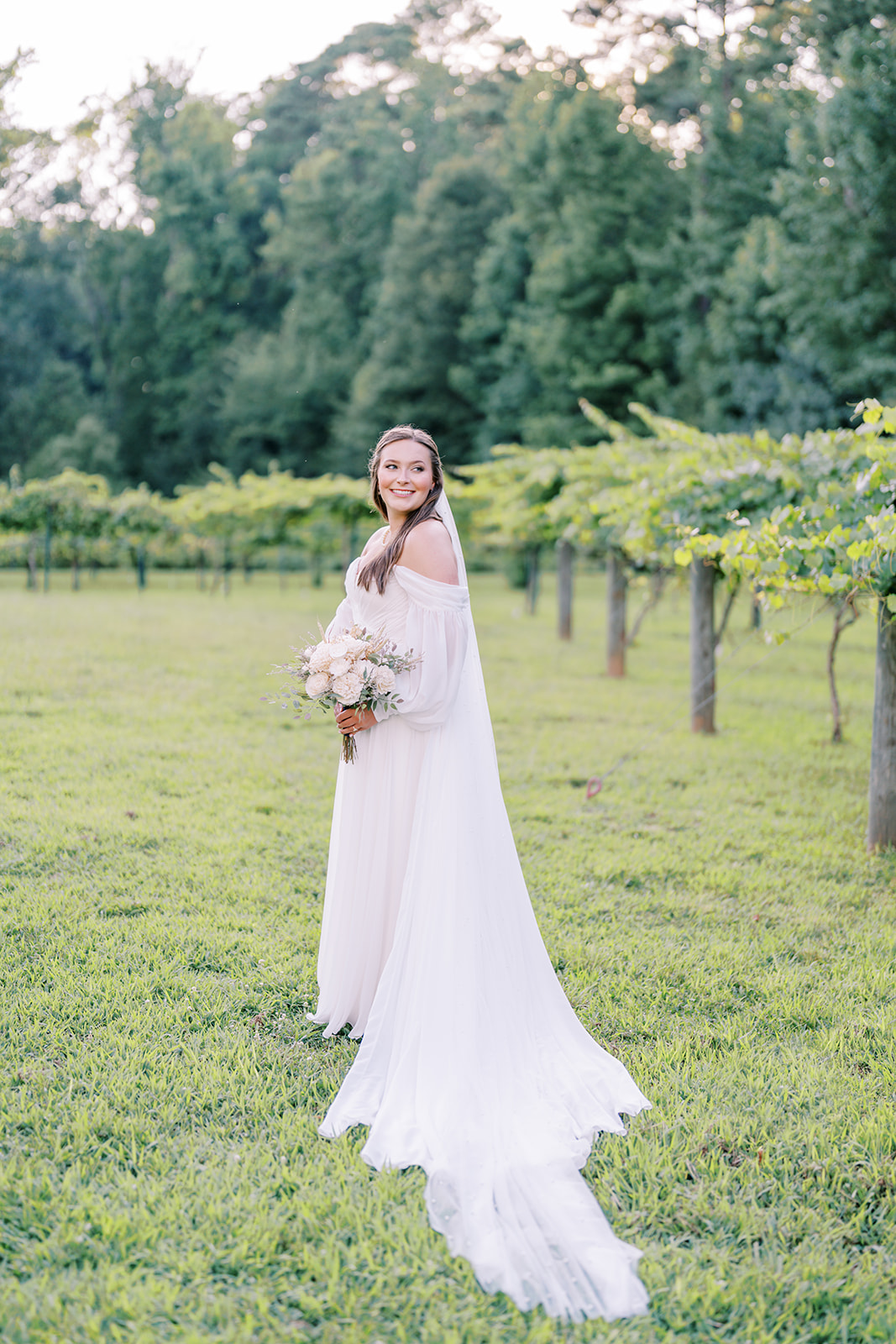 Cameron looks elegantly over her shoulder as her veil trails behind her in the vineyard at The Vines of Lexington in South Carolina, captured by North Carolina wedding photographer Jessica Leigh Mason.