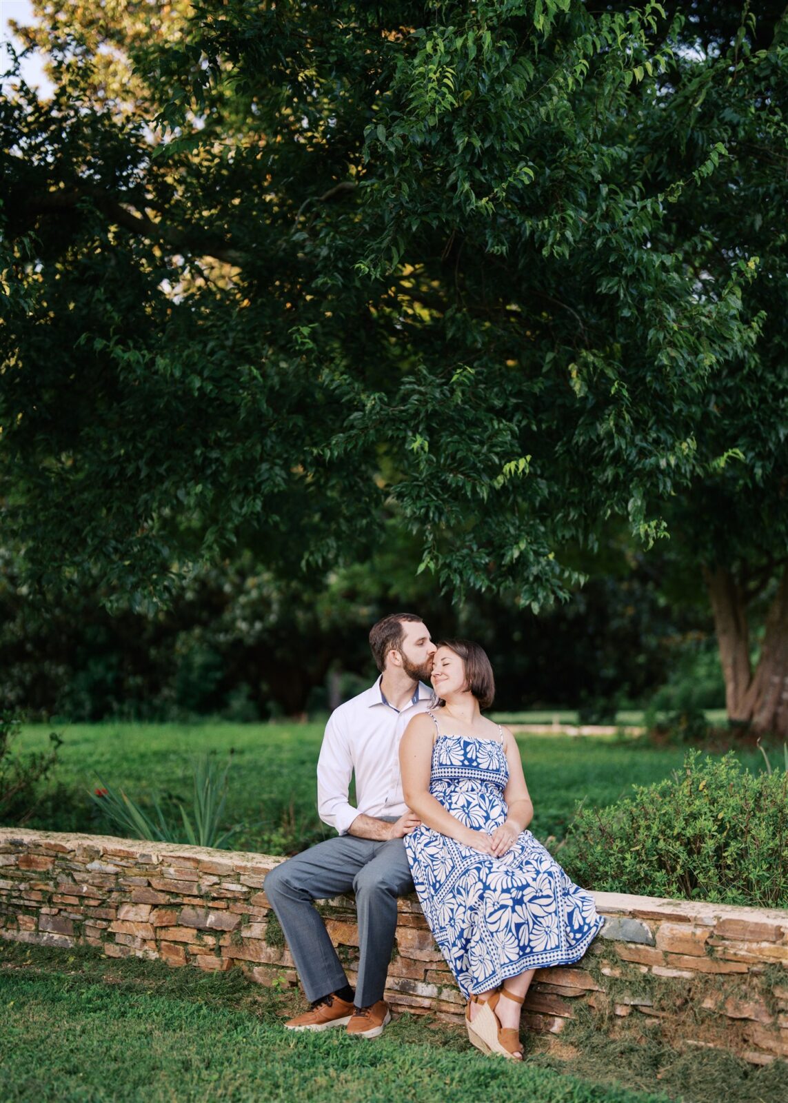 Katie and Sam sitting close together on a stone wall at Ayr Mount in Hillsborough NC, snuggling during their anniversary photo session.