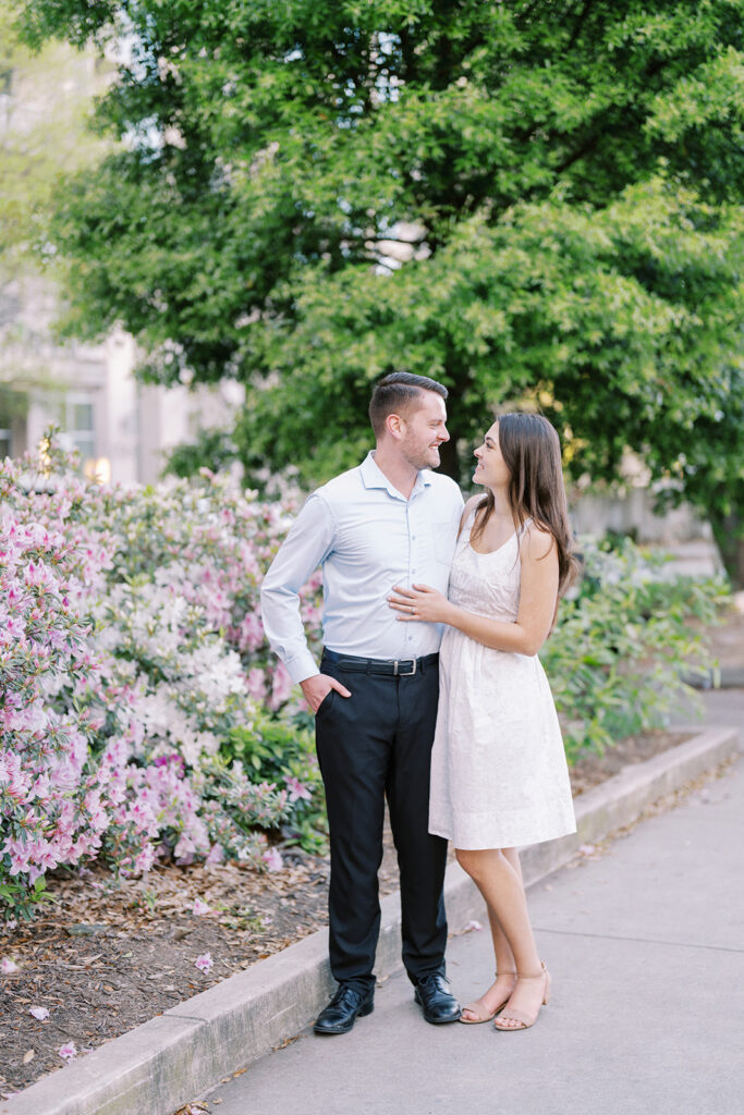 Full-length engagement portrait of Rachel and Nate standing in front of blooming azaleas at Romare Bearden Park in Charlotte, North Carolina