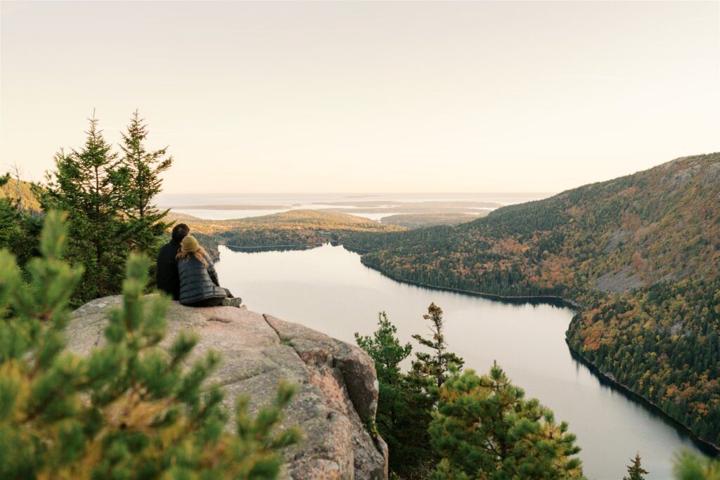 Engaged couple snuggled close at sunset on North Bubble overlooking Eagle Lake in Acadia National Park

