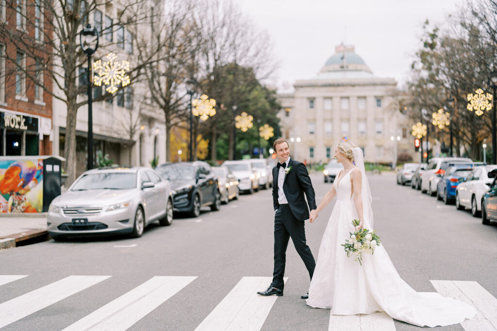 Bride and groom walking hand in hand across a street in downtown Raleigh near Stockroom 230