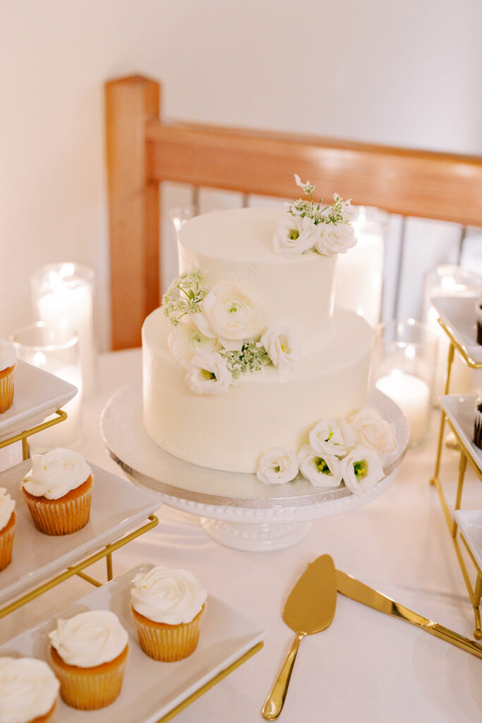 Candle-lit wedding cake surrounded by cupcakes at a reception dessert table