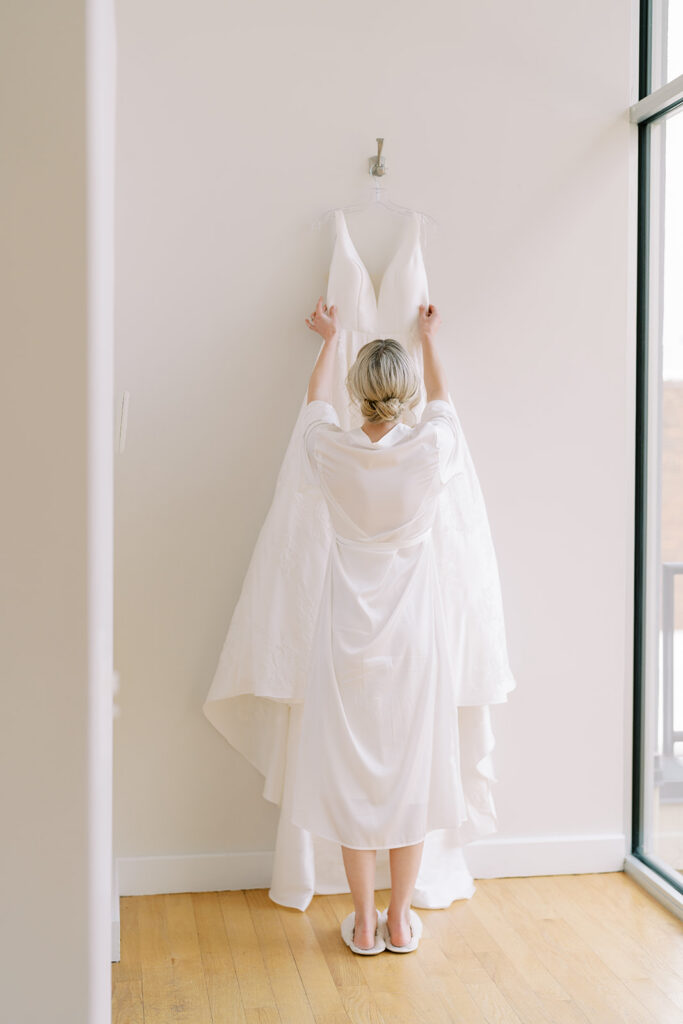 Bride reaching for her wedding dress in a softly lit bridal suite