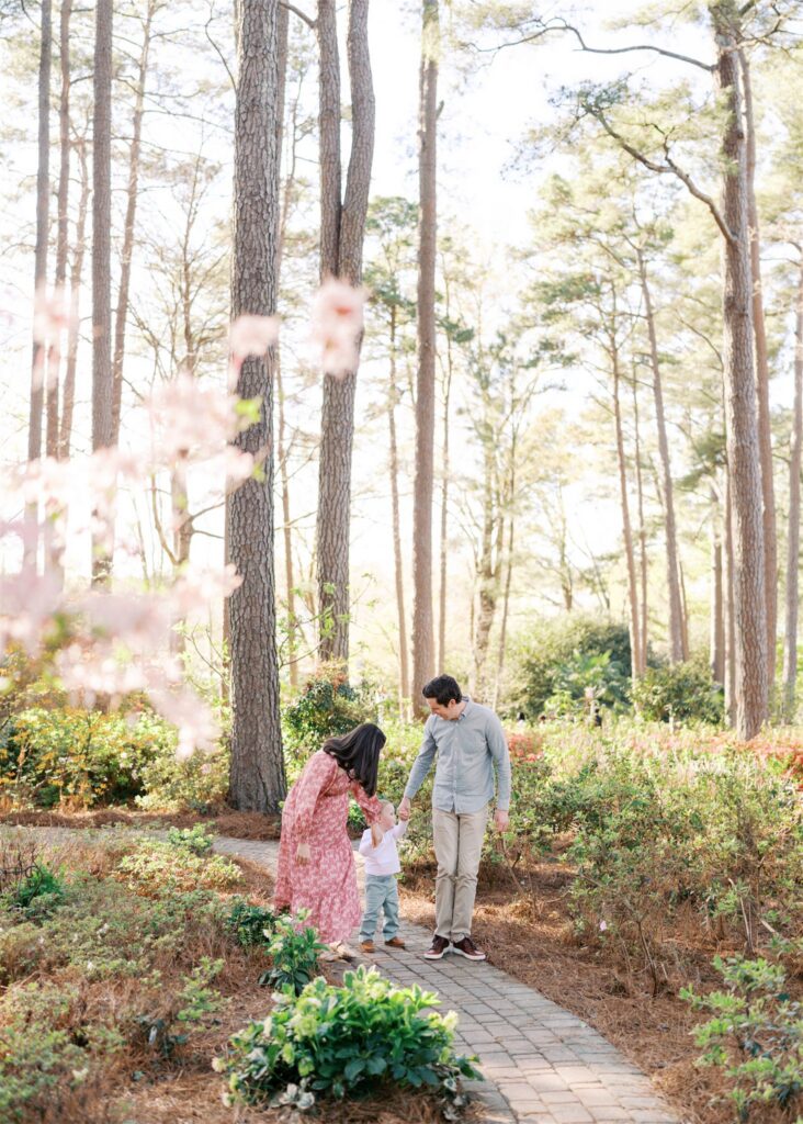 Candid shot of expecting family walking hand in hand through spring azalea blooms at WRAL Azalea Gardens Raleigh NC captured by Raleigh family photographer Jessica Leigh Photography