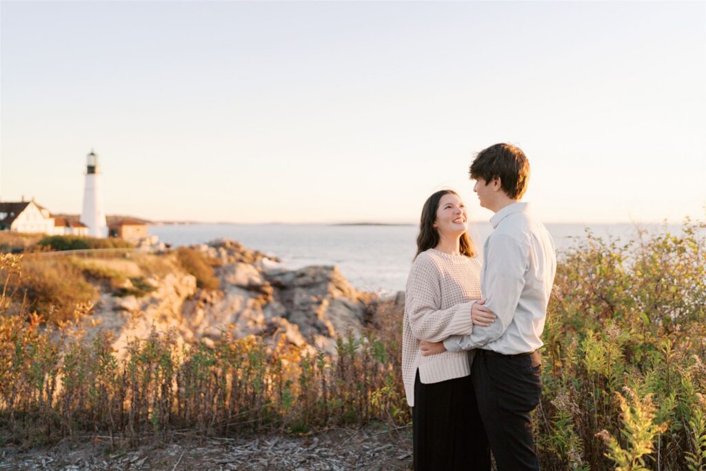 Couple photographed at sunrise during a Maine coast engagement session with Portland Head Light glowing in the golden morning light behind them