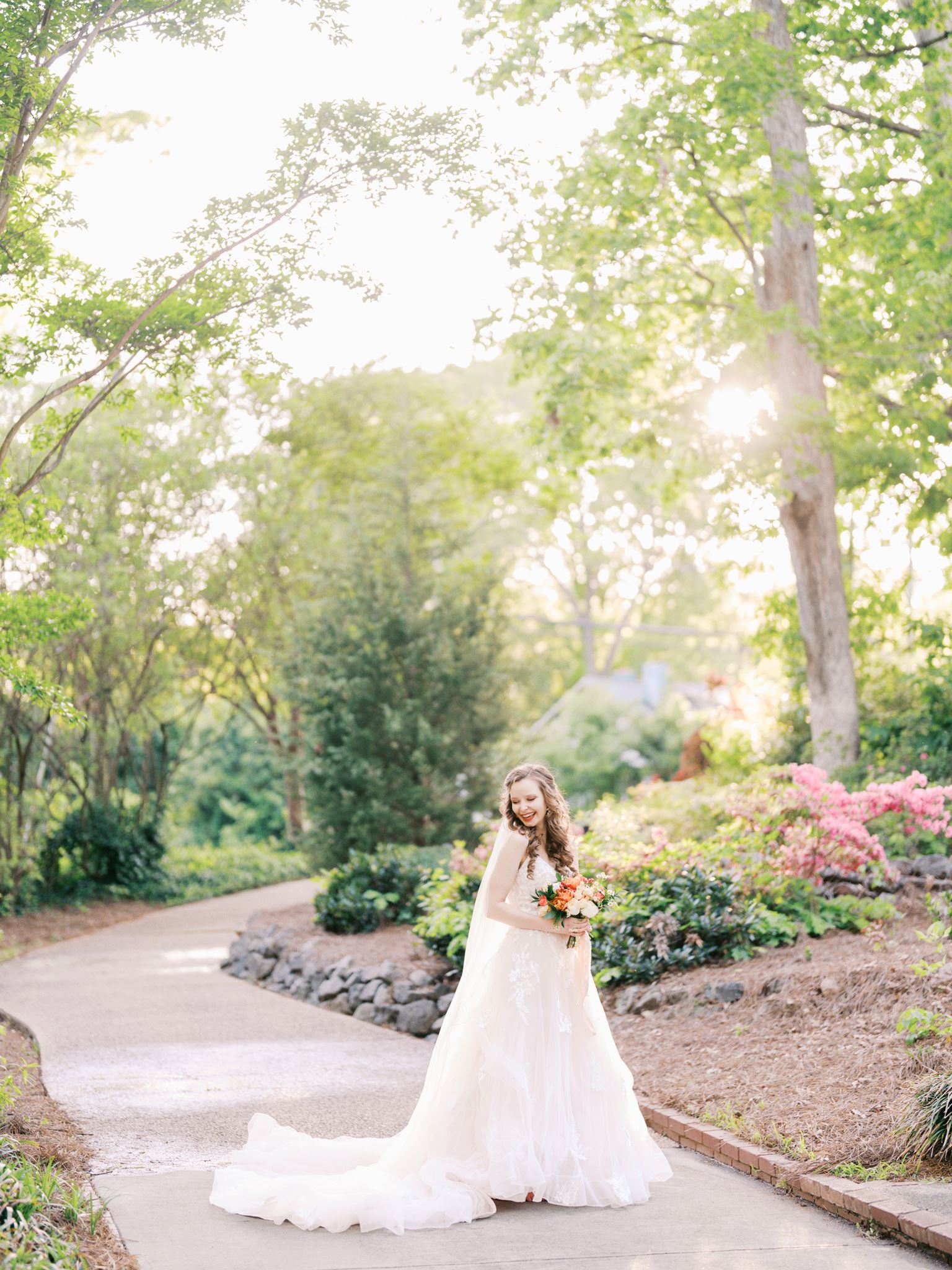 Full-length bridal portrait of a bride on a sunlit garden path surrounded by blooming flowers and greenery