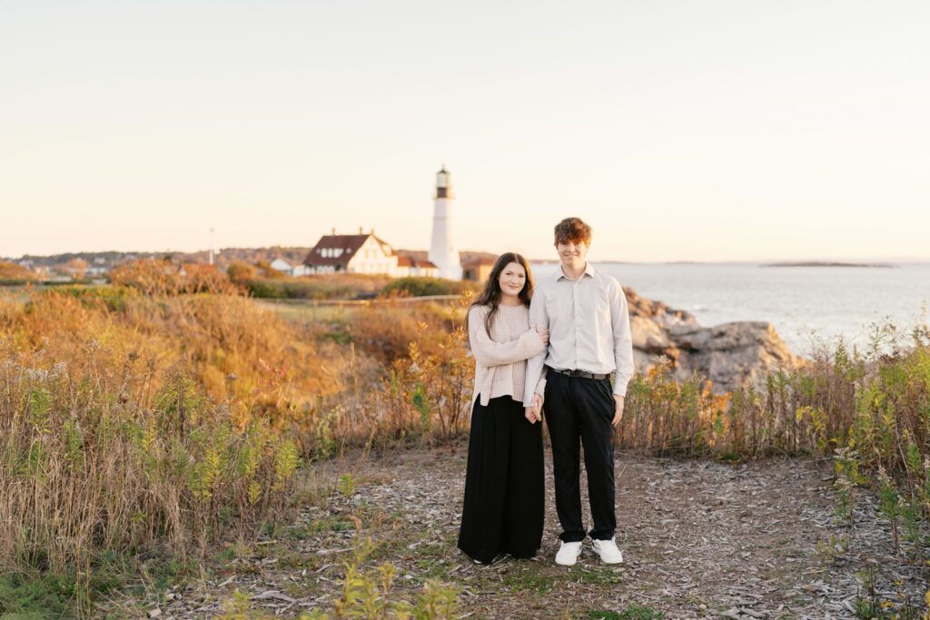 Full-length portrait of an engaged couple playing in a grassy field near Portland Head Light at sunrise
