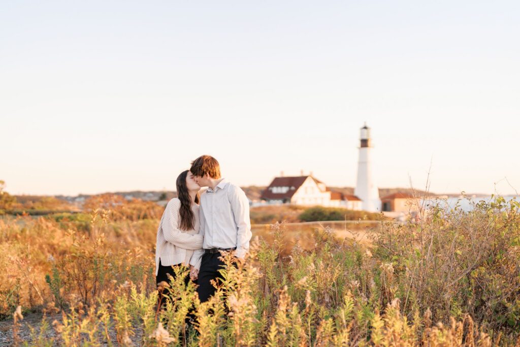 Couple sharing a soft kiss after running through a grassy field during a Portland Head Light engagement session at sunrise