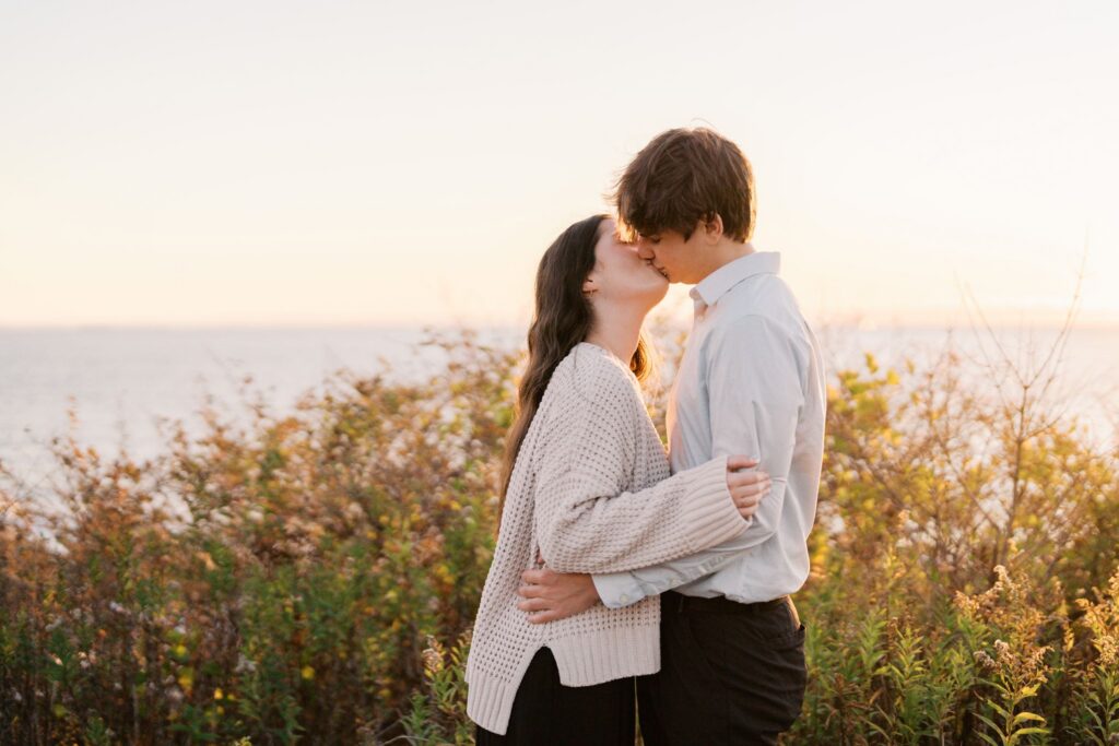 Close-up engagement portrait of a couple holding each other with warm coastal light and water in the background