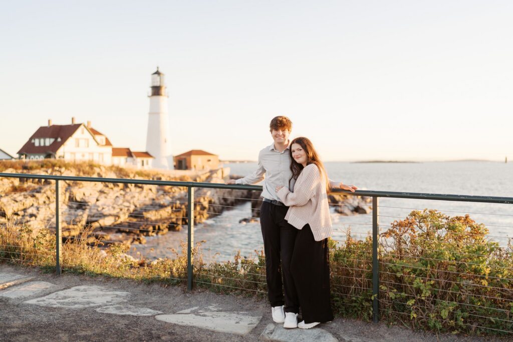 Couple leaning on a railing and looking toward the camera during a Portland Head Light engagement session in Cape Elizabeth, Maine