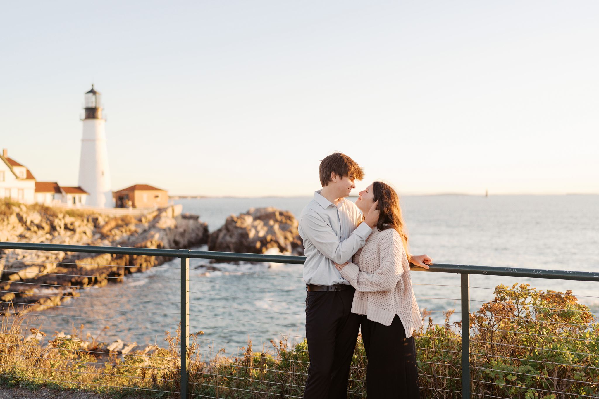 Engaged couple posing near the lighthouse with the Maine coastline behind them at sunrise