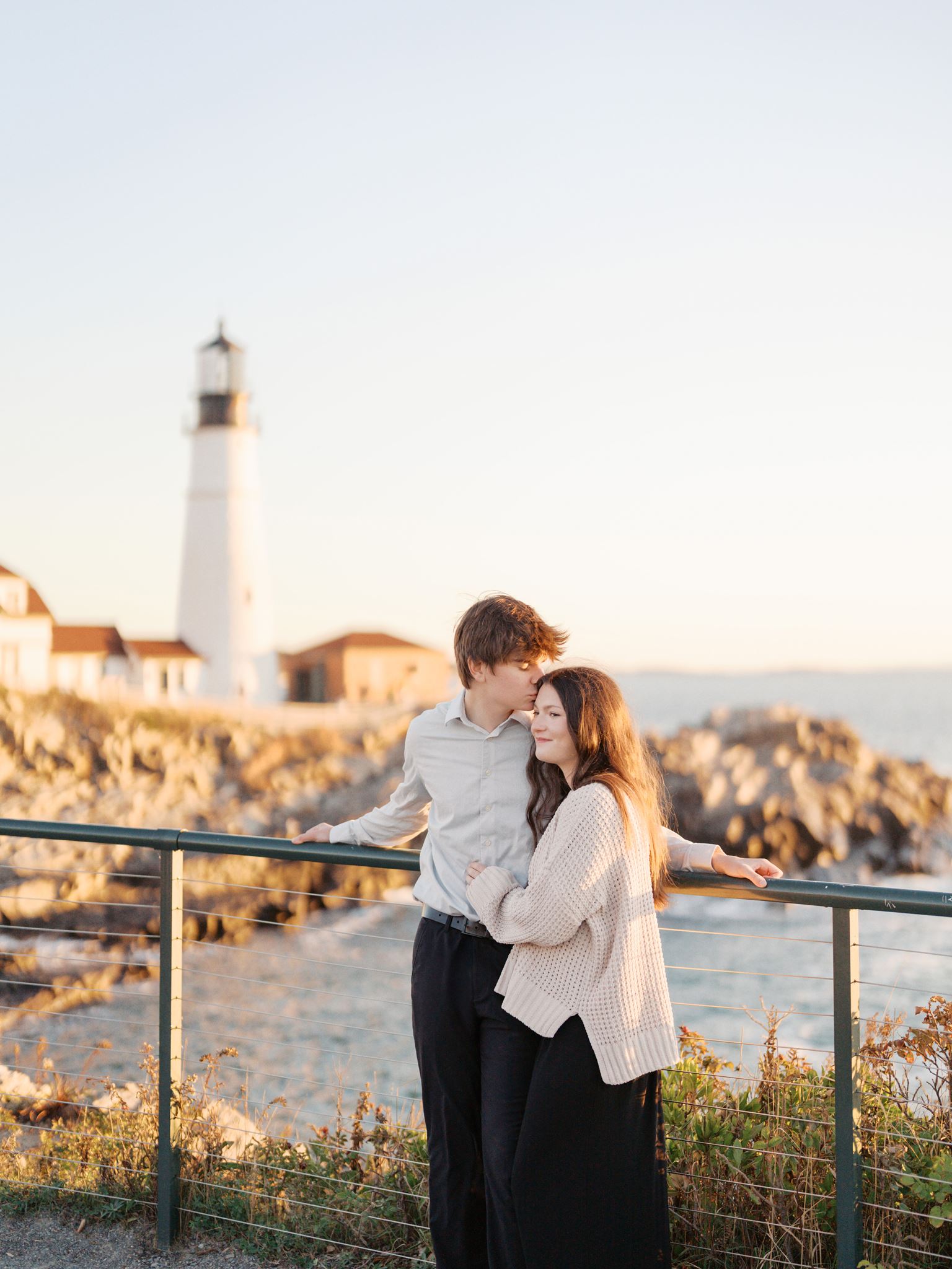 High school sweethearts looking at the camera with the iconic Portland Head Light behind them