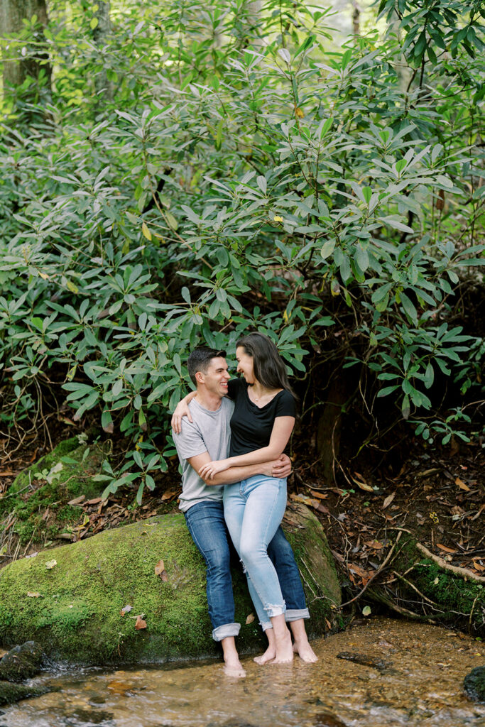 Engaged couple sitting barefoot with their feet in the creek at their Blue Ridge Parkway engagement session near Boone, NC